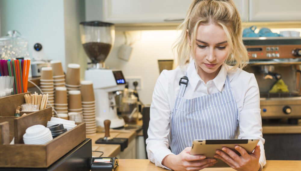 blonde-young-woman-standing-coffee-shop-counter-looking-digital-tablet